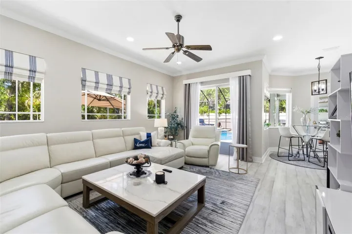 Bright and comfortable living room featuring a spacious sectional sofa, perfect for relaxing or entertaining. Large windows fill the space with natural light and offer pleasant views of the surrounding greenery. The room is complemented by neutral tones, modern finishes, and durable wood-look flooring, creating a clean and inviting atmosphere.