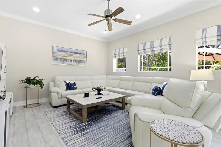 Bright and comfortable living room featuring a spacious sectional sofa, perfect for relaxing or entertaining. Large windows fill the space with natural light and offer pleasant views of the surrounding greenery. The room is complemented by neutral tones, modern finishes, and durable wood-look flooring, creating a clean and inviting atmosphere.