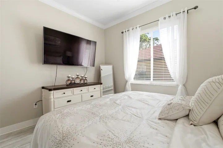 Light and airy bedroom featuring a soft neutral palette and elegant finishes throughout. Large windows with sheer curtains allow for abundant natural light, creating a warm and inviting atmosphere. The room offers a comfortable layout with stylish furnishings, ample space, and a serene feel—perfect for guests or family living.