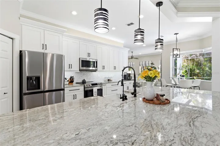 Stunning opBeautifully designed open-concept living space showcasing a spacious chef’s kitchen with a large granite island, ample seating, and abundant cabinetry. Stainless steel appliances, modern pendant lighting, and elegant finishes create a clean, upscale feel. The layout flows seamlessly into the living and dining areas, with sliding glass doors opening to the pool area—perfect for indoor-outdoor entertaining and everyday living.en-concept kitchen designed for both everyday living and entertaining, featuring a large center island with farmhouse sink, granite countertops, and ample cabinetry. Modern pendant lighting, stainless steel appliances, and sleek finishes create a clean, contemporary look. The space flows seamlessly into a bright dining area with large windows overlooking the pool and lush landscaping, while sliding glass doors provide easy indoor-outdoor living.