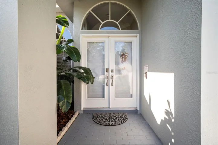 Inviting front entry featuring elegant double doors with decorative glass inserts and an arched transom window that adds architectural charm and natural light. The covered entryway offers a welcoming first impression, complemented by tropical landscaping and a clean, well-maintained exterior.