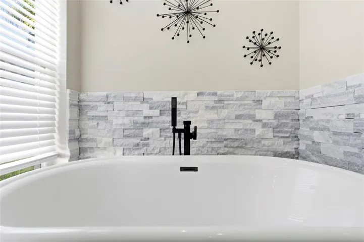 Elegant freestanding soaking tub set against a designer stone accent wall, creating a true spa-like retreat. Sleek matte black fixtures add a modern touch, while nearby windows provide natural light for a bright, relaxing atmosphere. Perfect for unwinding, this beautifully appointed space blends comfort and contemporary style.