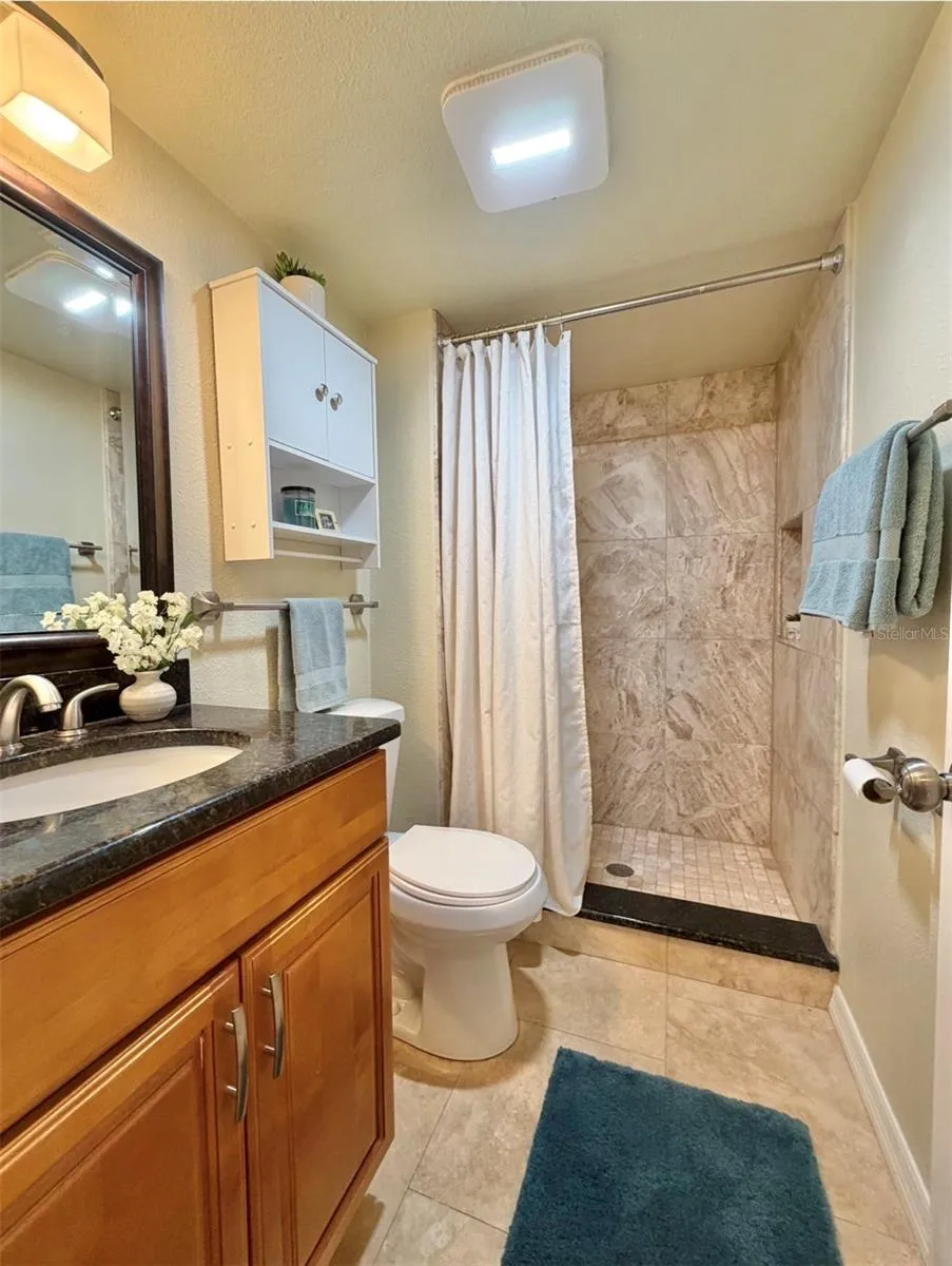 The second bathroom features luxurious travertine floors, a floor-to-ceiling tiled walk-in shower with a niche, granite countertops, a storage vanity, new satin nickel fixtures, a storage cabinet above the toilet, and updated lighting.
