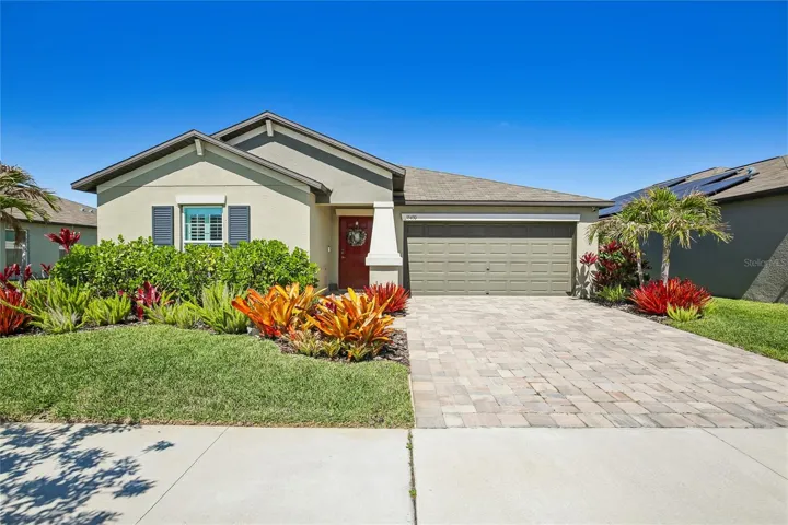 Single-story stucco home with a pavered driveway, two-car garage, and a bold red front door. Mature tropical landscaping with colorful bromeliads and palms frames the entry