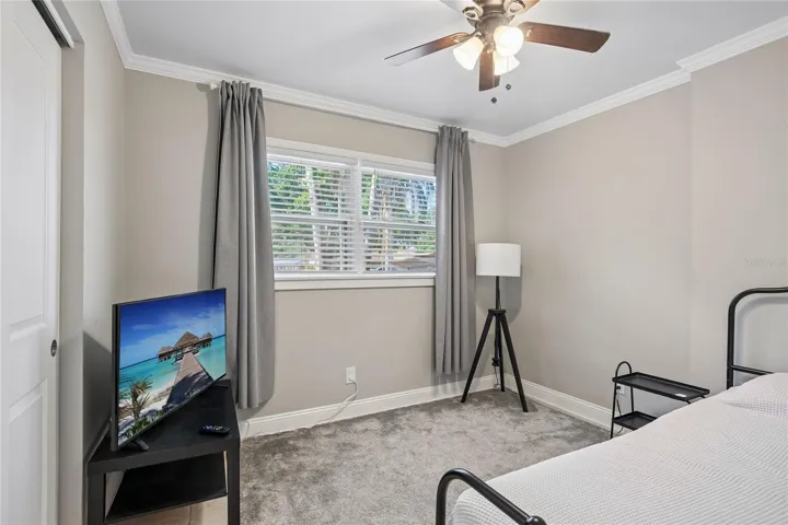 Bedroom Two – Bedroom two offers a comfortable layout with crown molding, a ceiling fan, and a wide window with blinds and curtains. Neutral finishes and soft carpeting create a clean, flexible space.