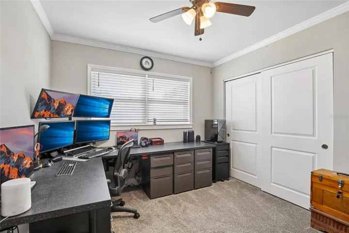 Bedroom Three – Bedroom three is currently arranged as a home office, featuring plush carpeting, crown molding, a ceiling fan, and a wide window with blinds. The room offers flexible use as a bedroom, workspace, or hobby area.