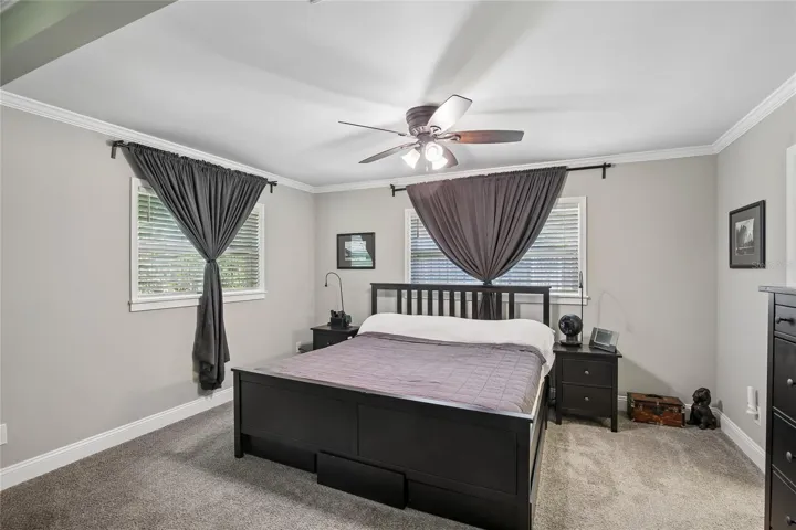 Primary Bedroom – The primary bedroom offers a spacious layout with plush carpeting, crown molding, a ceiling fan, and multiple windows with blinds and curtains. Neutral finishes create a calm, comfortable retreat.