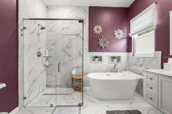 The spa-inspired primary bath showcases dramatic floor-to-ceiling designer tile, a frameless glass walk-in shower with rain shower head, freestanding soaking tub, designer quartz countertops, and upgraded plumbing fixtures. Decorative wall niches, abundant natural light, and contemporary finishes create a luxurious retreat-like atmosphere.