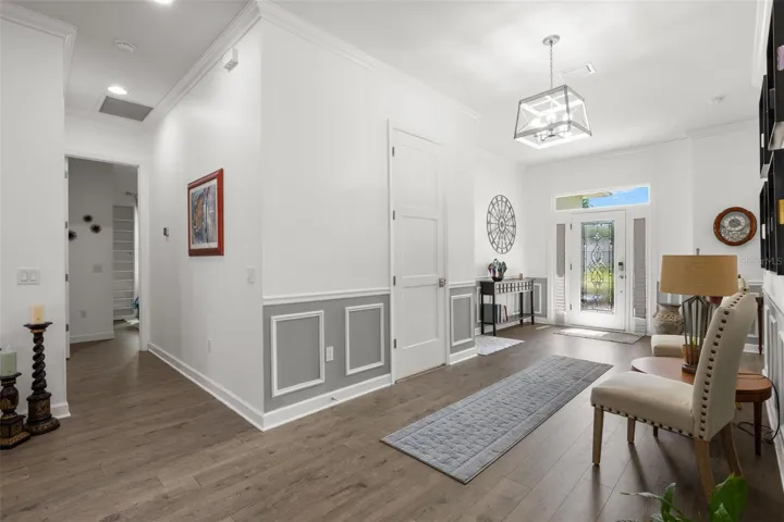 The oversized foyer is framed by extensive crown molding, oversized base trim, wainscoting, chair rail detailing, recessed lighting, and wide-plank luxury vinyl flooring. A statement chandelier, elegant leaded glass entry door with sidelights and transom window, and oversized paneled interior doors contribute to the home’s polished architectural character.