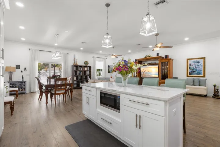 The oversized center island serves as a striking focal point within the open-concept living space, offering designer quartz countertops, additional storage, integrated 2nd microwave, pendant lighting, and generous seating. Open sightlines connect the kitchen, dining area, and great room beneath high ceilings with recessed lighting and dual contemporary ceiling fans.