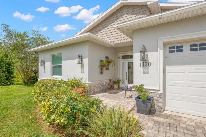 The covered front entry combines stacked stone accents, brick pavers, decorative lighting, and architectural detailing for a polished first impression. A leaded glass front door with sidelights welcomes guests into the home, while manicured landscaping and rain gutters add both beauty and functionality.