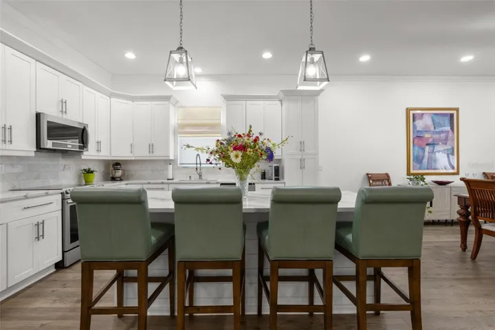 The oversized center island provides extensive prep space, additional storage, integrated microwave, and comfortable seating beneath contemporary pendant lighting. Crisp white shaker cabinetry with crown molding, designer quartz countertops, upgraded stainless appliances, recessed lighting, and an elegant onyx marble backsplash create a clean, sophisticated kitchen design.