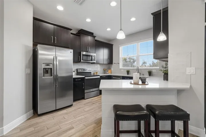 Updated kitchen featuring sleek white quartz countertops, stainless steel appliances, dark shaker-style cabinetry, recessed lighting, and a spacious breakfast bar perfect for casual dining and entertaining. Large windows bring in natural light, while the modern backsplash and wood-look tile flooring add warmth and style throughout