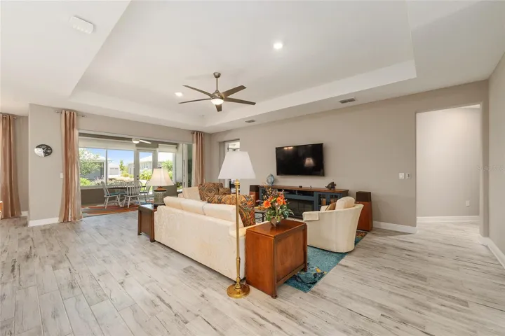 Spacious great room with tray ceiling, recessed lighting, ceiling fan with light, ceramic plank flooring, and stacking sliding doors opening to the enclosed lanai.