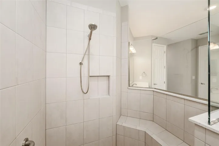 First primary bathroom ensuite with zero-entry walk-in shower with built-in seating, glass enclosure, tile surround, niche, and handheld shower fixture.