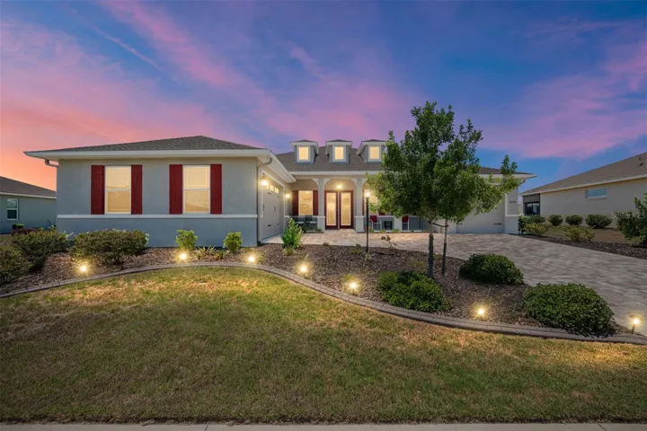 Stunning twilight view showcasing the home’s curb appeal, professional landscape lighting, paver driveway, covered front porch, and inviting lead glass double-door entry.