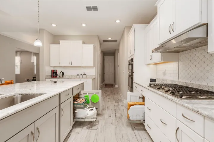Bright kitchen workspace with a large single-basin sink, built-in soap dispenser, pull-out drawers, neutral backsplash, Cambria Quartz island, and open flow to the main living area.