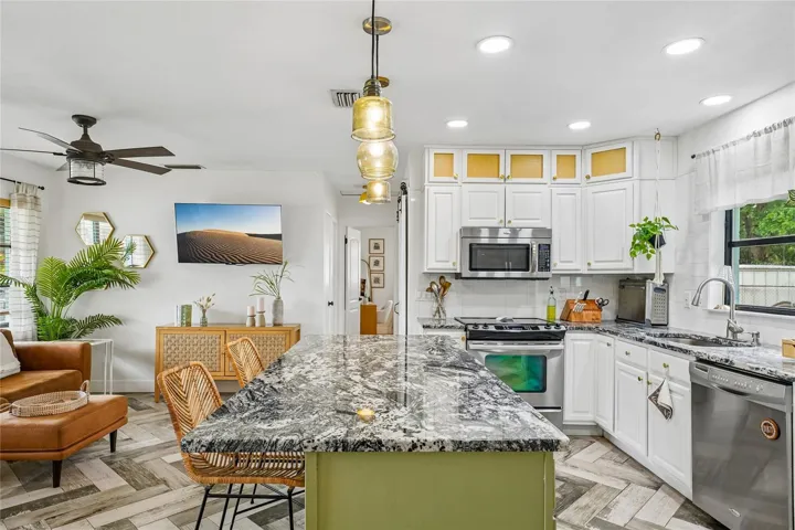 Kitchen — View from the island looking toward the cooking area, with white cabinetry, subway tile backsplash, stainless steel range and dishwasher, and dark granite countertops. Open sightline to the living room beyond, with herringbone tile flooring throughout.