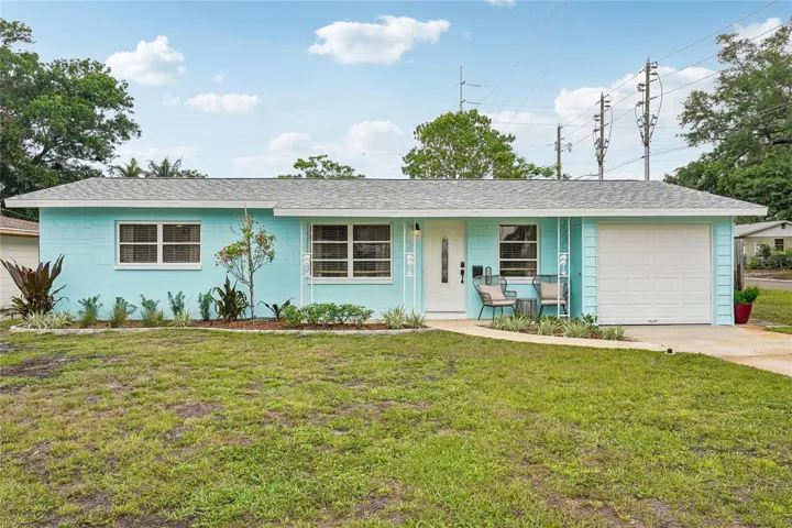 Front Exterior — Concrete block ranch-style home with a fresh aqua exterior, white garage door, and covered front entry with seating area. Mature landscaping, tropical plantings, and a lush front lawn add to the inviting curb appeal.