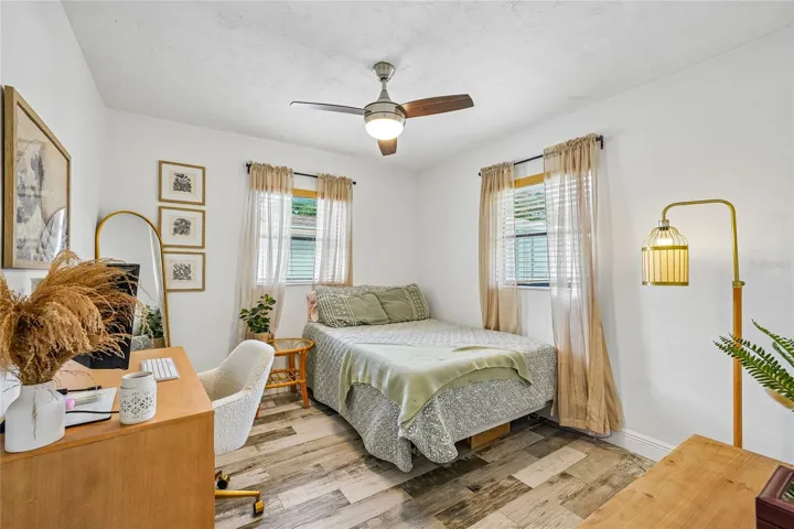 Bedroom 1 — Bedroom with tile flooring, ceiling fan, and two windows with blinds and sheer curtains. Room accommodates a queen bed and desk area.