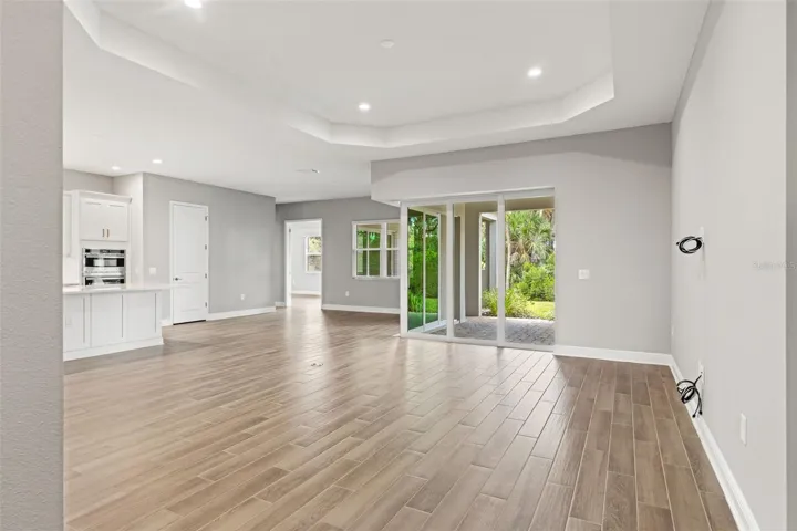 Expansive open-concept great room with tray ceilings, wood-look tile flooring, and oversized sliding glass doors that bring in natural light and create seamless indoor-outdoor flow.