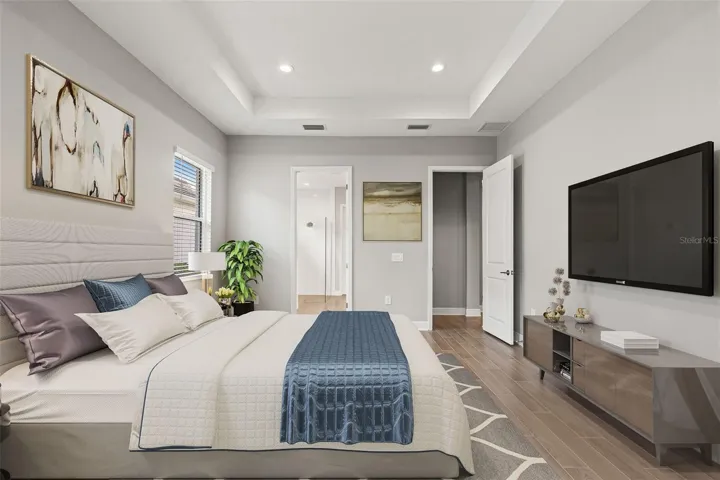 Spacious primary suite featuring tray ceilings, wood-look tile flooring, and a relaxing atmosphere with ample room for a sitting area and media setup.
