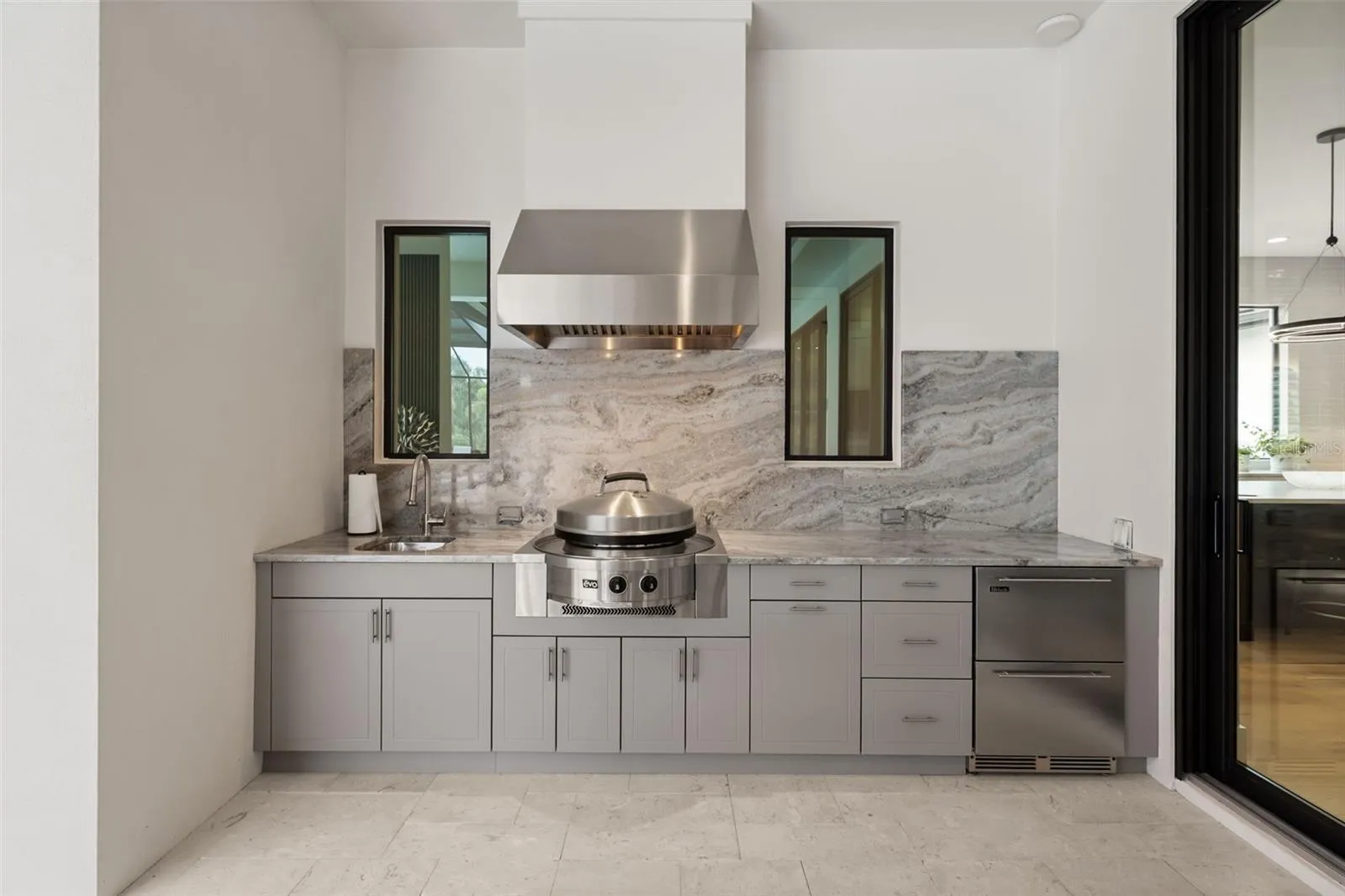 The fully equipped outdoor kitchen rivals the interior kitchen in both style and function, featuring an Evo gas grill, sink, dual Perlick refrigerator drawers, custom outdoor cabinetry with integrated trash storage, granite countertops and backsplash.