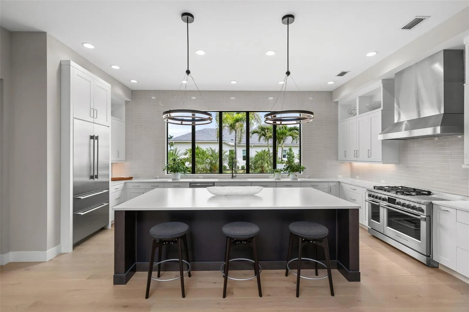 Matte-finished quartz countertops, a large center island with seating, tile backsplash, wood cabinetry with display shelving and pendant lighting create a polished and inviting atmosphere.