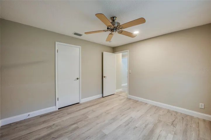 Bright bedroom showcasing vinyl flooring, neutral beige walls, a classic ceiling fan, and access to both a closet and the hallway.