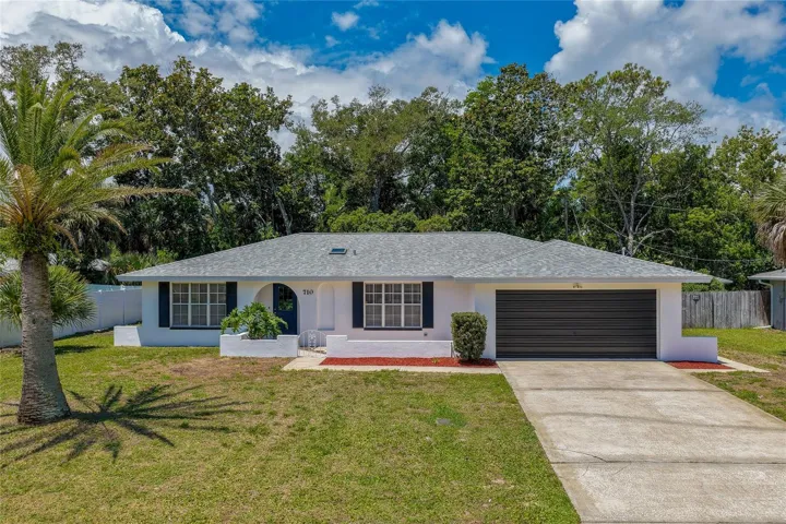 Charming ranch home featuring a fresh white stucco exterior, contrasting black shutters, and an attached two-car garage. The concrete driveway leads up to a neat front yard bordered by mature trees.