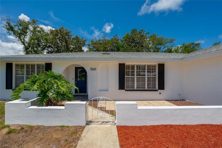 The welcoming front entry features an arched alcove, framed by classic black shutters and crisp white walls. A low courtyard wall and decorative iron gate enhance the home's distinctive curb appeal.