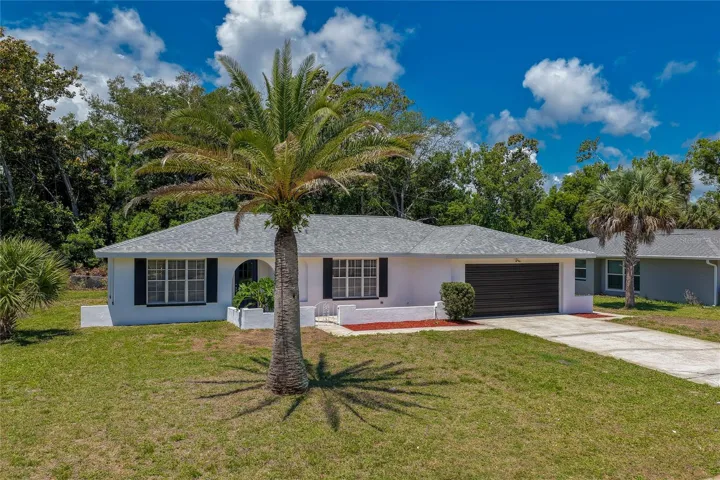 A wide exterior perspective highlighting the property’s expansive front lot and vibrant tropical landscaping. The architectural roofline and clean white brick details blend seamlessly into the quiet, established neighborhood.