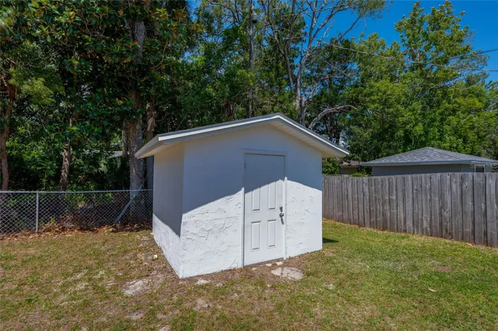 A standalone stucco storage shed features a sturdy paneled door and matching shingled roof, offering secure additional storage. It sits tucked away in the corner of the yard against a backdrop of mature, shady trees.