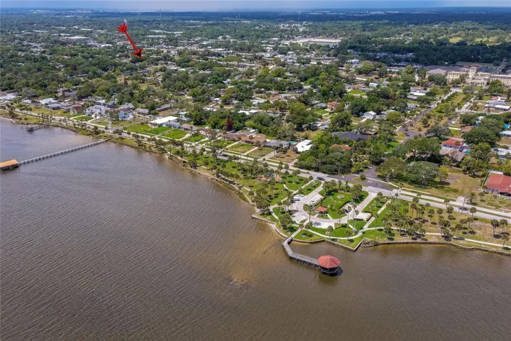 This beautiful waterfront aerial highlights a public park featuring a gazebo, walking paths, and a pier extending into the water. A red arrow in the distance indicates the property's location within the nearby coastal community.