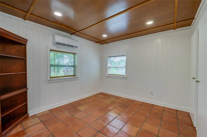 A versatile bedroom boasting warm tile flooring, crisp white wood-paneled walls, and a unique wood-paneled ceiling with recessed lighting. The space is equipped with a wall-mounted mini-split AC unit and includes a built-in wooden bookcase.
