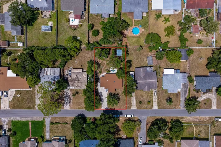 A top-down aerial view highlights a residential lot outlined in red, showing a brown-roofed home surrounded by lush green trees and an expansive driveway. The surrounding neighborhood is dotted with single-family homes, manicured lawns, and backyard pools.
