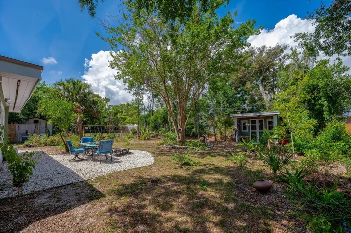 An expansive backyard oasis featuring a gravel patio area complete with outdoor seating under a canopy of mature trees. A charming detached storage shed sits nestled among lush greenery, offering plenty of space for a relaxed, nature-filled coastal lifestyle.
