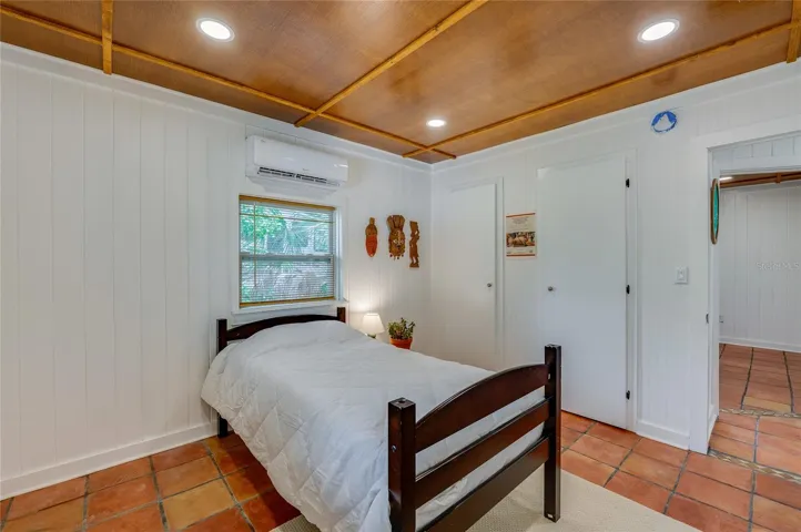 Alternate view of the bedroom highlighting the warm wood ceiling, and a doorway leading into the hallway. A mini-split unit ensures comfort above the window.