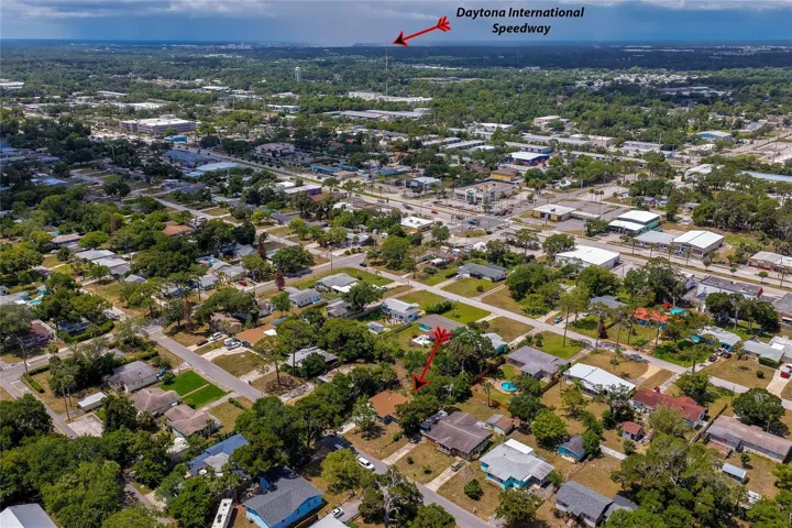 An elevated aerial view shows the property nestled within a quiet neighborhood grid, with a red arrow pinpointing the house. The far horizon highlights the distant location of the Daytona International Speedway beneath a dramatic sky.