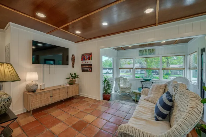 Cozy conversational layout looking from the living area toward the bright sunroom, seamlessly connecting the two spaces. This viewpoint emphasizes the continuity of the handsome terracotta flooring and paneled wood ceilings.