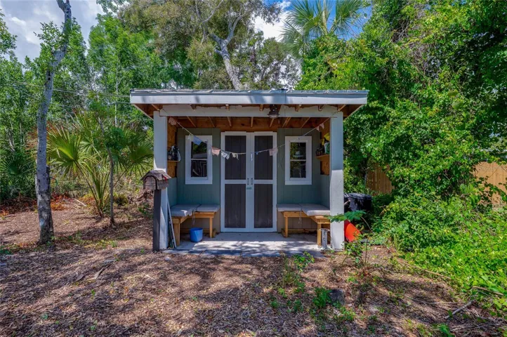 Charming detached storage shed or workshop featuring built-in bench seating, double doors, and a shaded front overhang surrounded by lush greenery.
