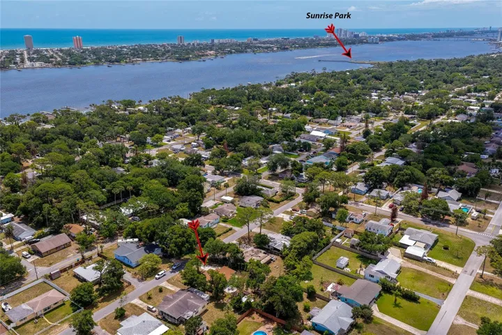 This scenic aerial shot emphasizes the property's proximity to a large body of water, with a red arrow pointing out the home. In the distance, a red indicator points across the water toward Sunrise Park and the coastal skyline.