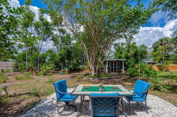 Inviting outdoor dining area featuring a table and blue chairs set on a white gravel patio, surrounded by a spacious, tree-lined yard. A charming green garden shed stands in the background under a bright, cloud-filled sky.