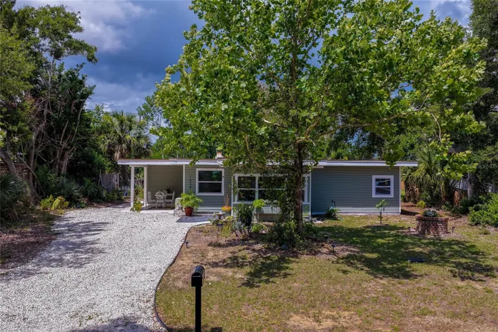 Charming single-story home nestled beneath a majestic shade tree, featuring a welcoming covered entry, lush tropical landscaping, and a spacious gravel driveway in a peaceful natural setting.