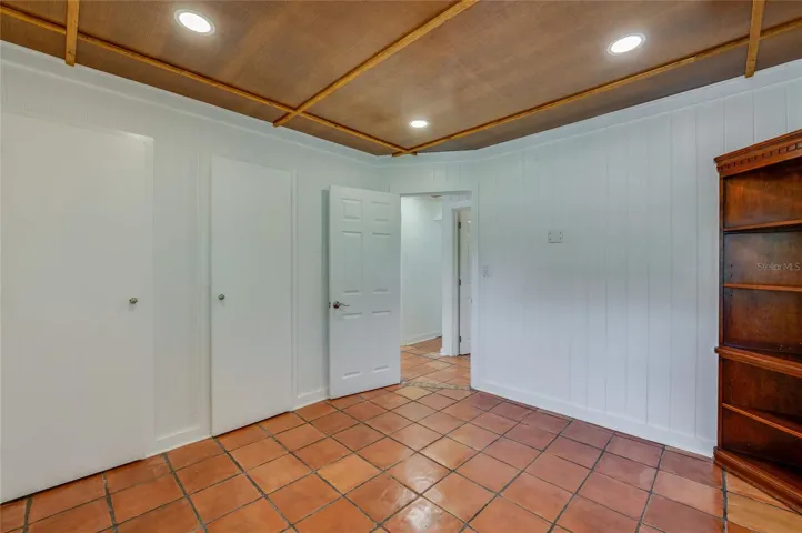 A bedroom featuring warm tile flooring, crisp white vertical paneled walls, and a rich wood-paneled ceiling with recessed lighting. A wooden corner bookshelf and multiple doorways anchor the space.