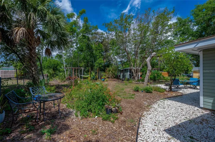 Lush, expansive backyard area offering a gravel patio space with outdoor seating nestled among mature trees and tropical greenery. A detached storage shed can be seen in the background, enhancing the functional outdoor living space.