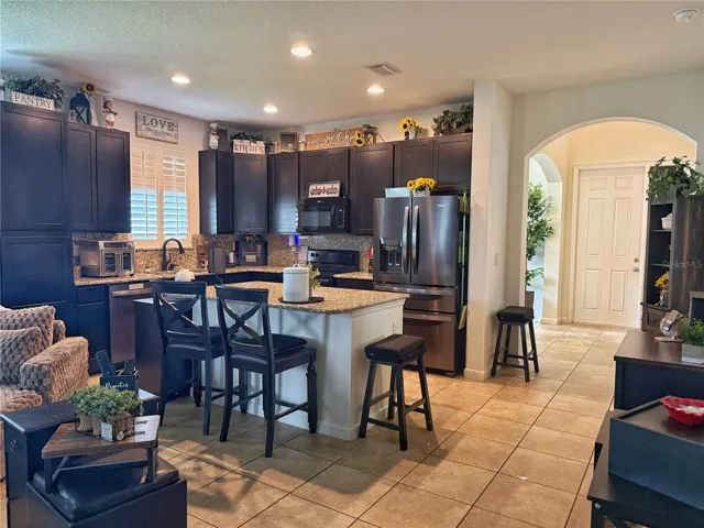 Kitchen with view of the Entry, ceramic Tile Flooring, kitchen island with bar, top seating, backsplash, stainless steel appliances, granite countertops, recessed, lighting, plantation shutters