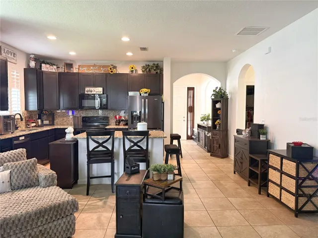 Additional view of kitchen from the living room with entry in the background, ceramic Tile Flooring, Tracie ceiling in the living room and recessed lighting in the kitchen, backsplash, island with bar top seating, 42 inch cabinets, stainless steel appliances, plantation shutters