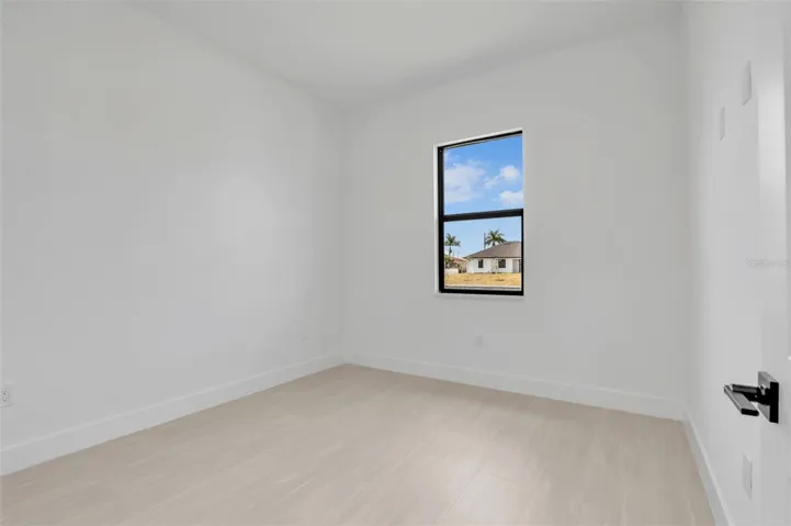 Guest Bedroom -Bright interior room featuring a single window, light wood-finish flooring, white baseboards, and a contemporary door handle