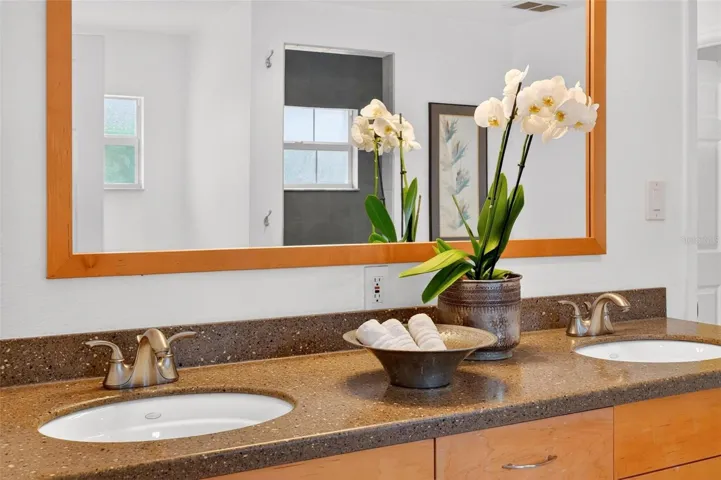 Spacious en-suite bath featuring a long vanity with dual sinks, warm wood cabinetry, and speckled solid-surface style countertops, offering generous prep and storage space.